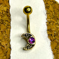 Gold-toned belly button ring with a crescent moon charm featuring an ornate filigree design and a purple gemstone, displayed on a white textured strip between cork backgrounds.