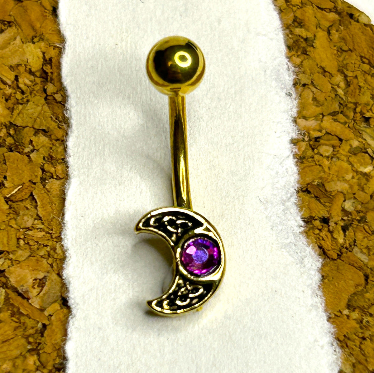 Gold-toned belly button ring with a crescent moon charm featuring an ornate filigree design and a purple gemstone, displayed on a white textured strip between cork backgrounds.