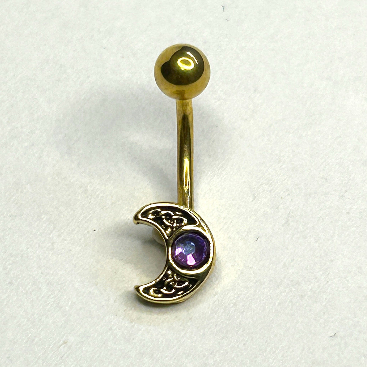 A gold-plated curved barbell belly button piercing with a crescent moon charm featuring an ornate filigree design and a small purple gemstone, shown on a plain white background.